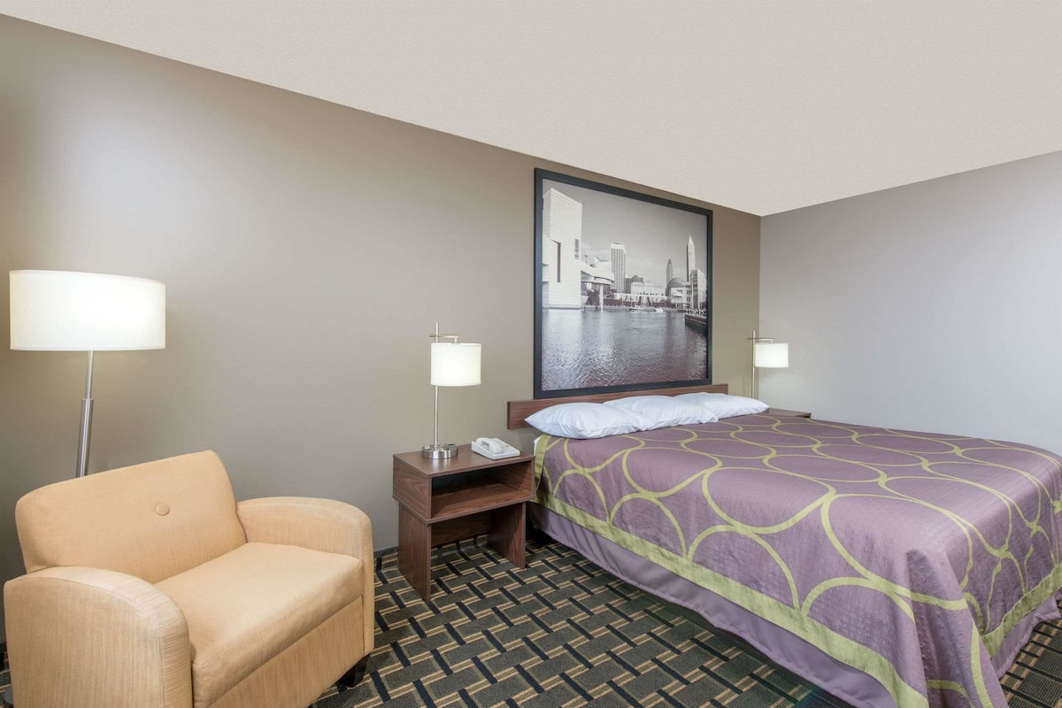 A hotel room with a bed, a chair, a nightstand, a lamp, and a cityscape painting on the wall.