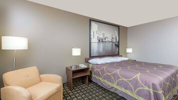 A hotel room with a bed, a chair, a nightstand, a lamp, and a cityscape painting on the wall.