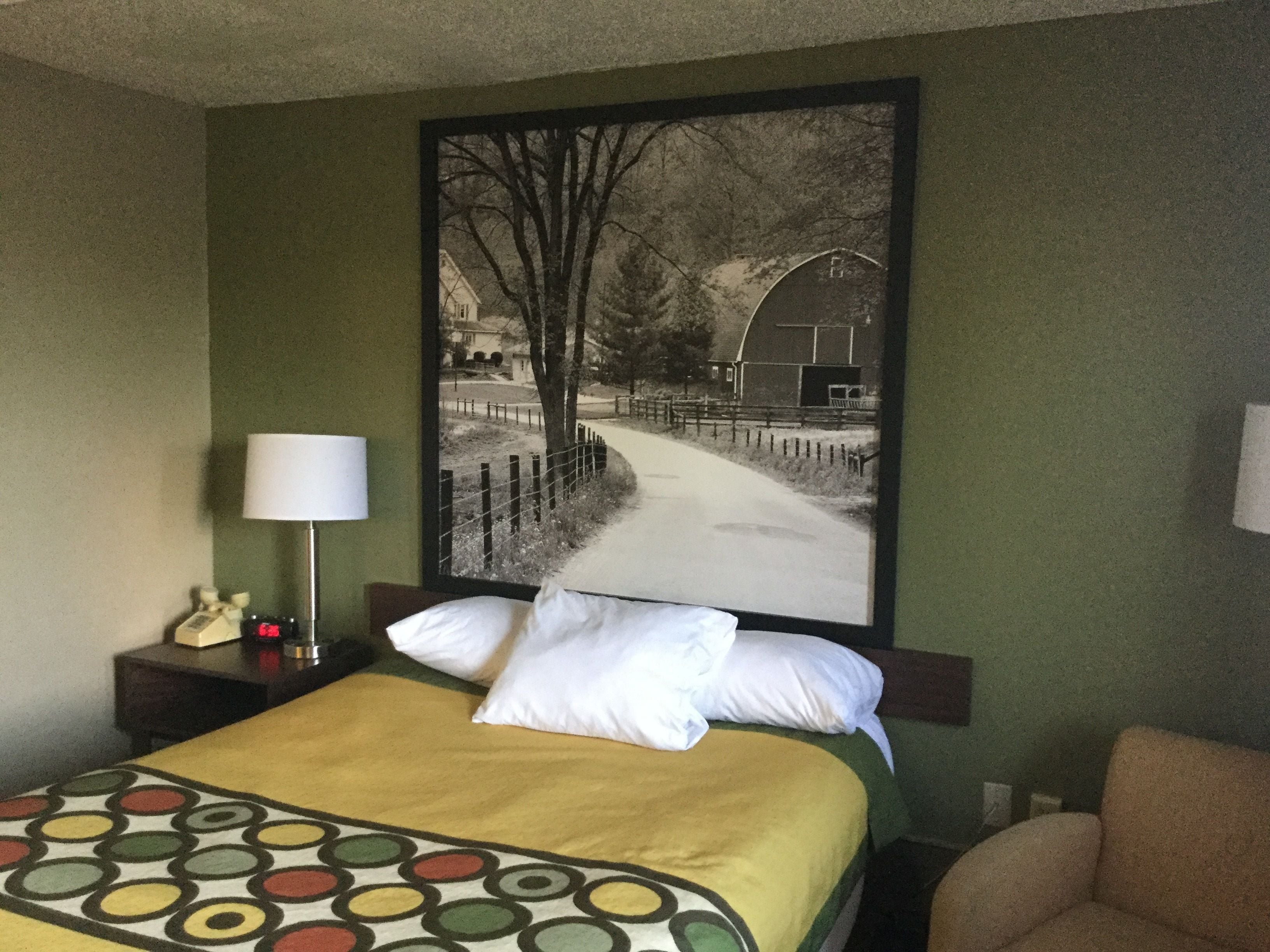 Ein Hotelzimmer mit einem Bett, einem Nachttisch, einer Lampe, einem Sessel und einem großen Bild in einem Rahmen an der Wand.