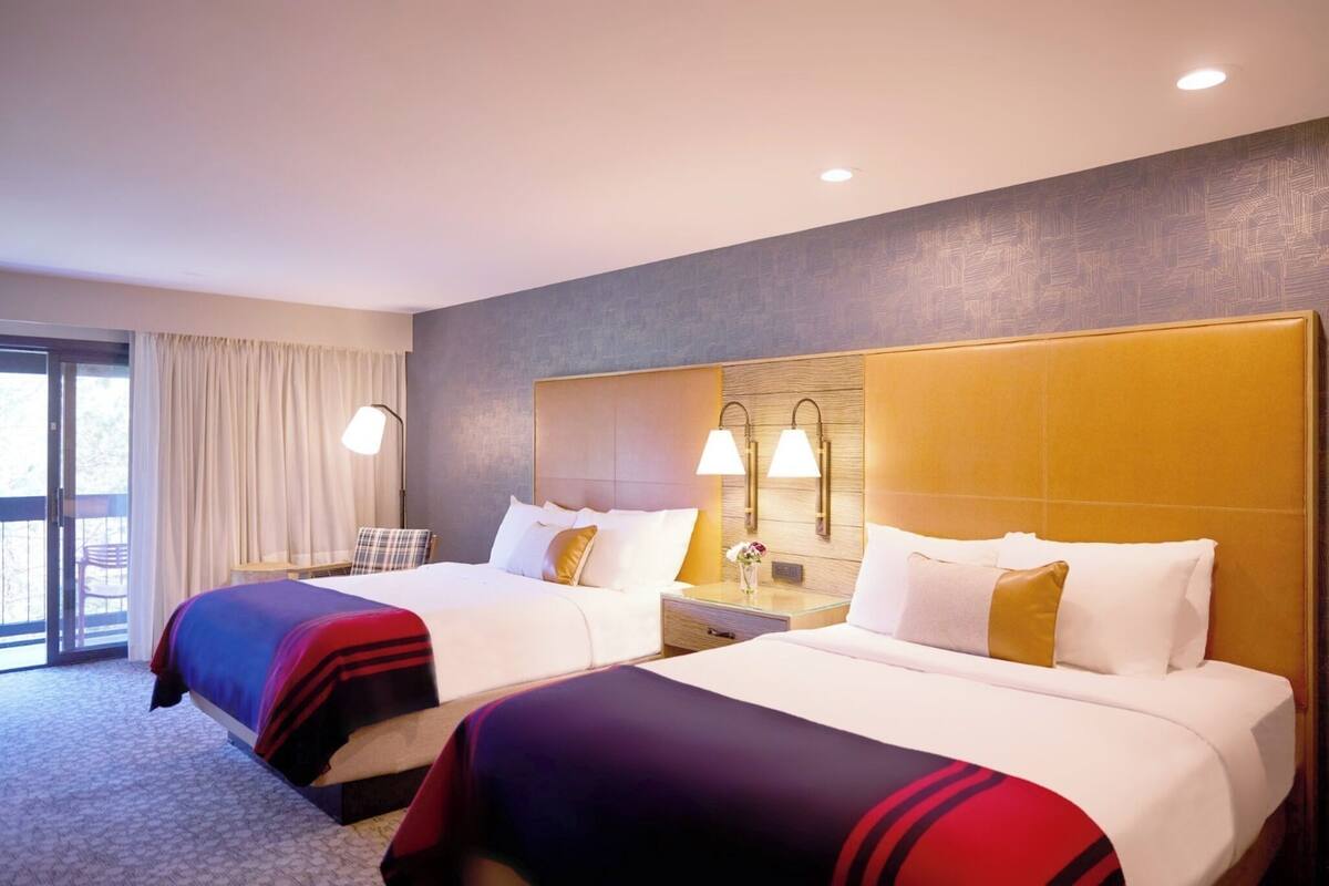 A hotel room with two beds, a large window with curtains, a nightstand with lamps, and a wall-mounted light fixture.