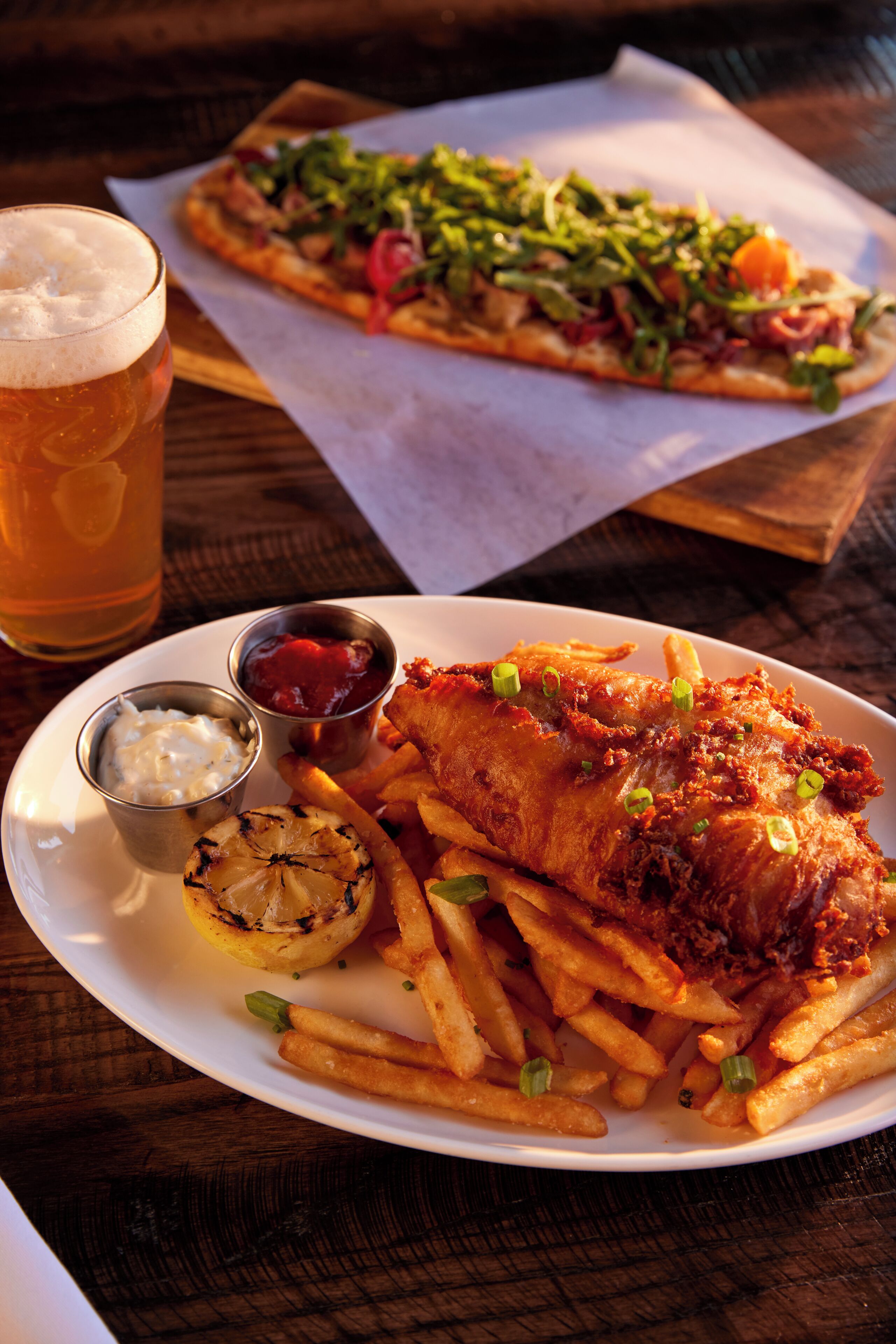 A plate of fish and chips with a side of fries, a small cup of ketchup, and a glass of beer on a wooden table.