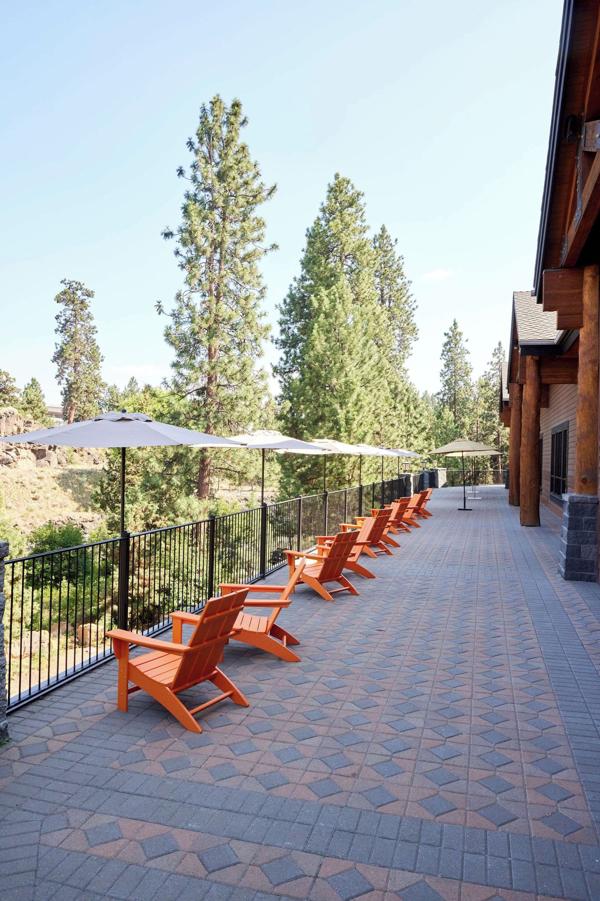 A covered outdoor seating area with chairs and umbrellas, surrounded by a wooden building and trees.