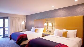 A hotel room with two beds, a large window with curtains, a nightstand with lamps, and a wall-mounted light fixture.