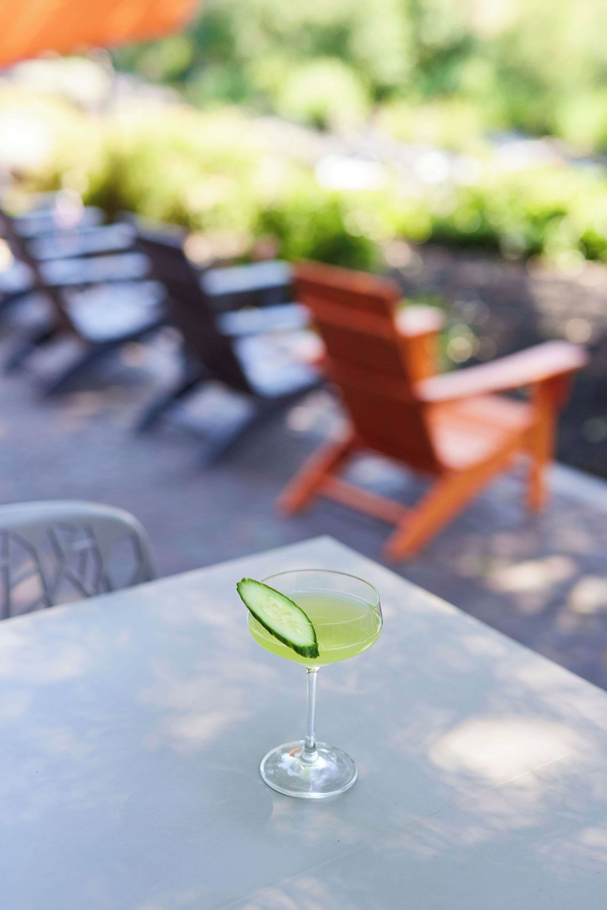 A table with a clear glass containing a green beverage, possibly a cocktail, with a slice of cucumber garnish. In the background, there are outdoor chairs and a blurred view of greenery.