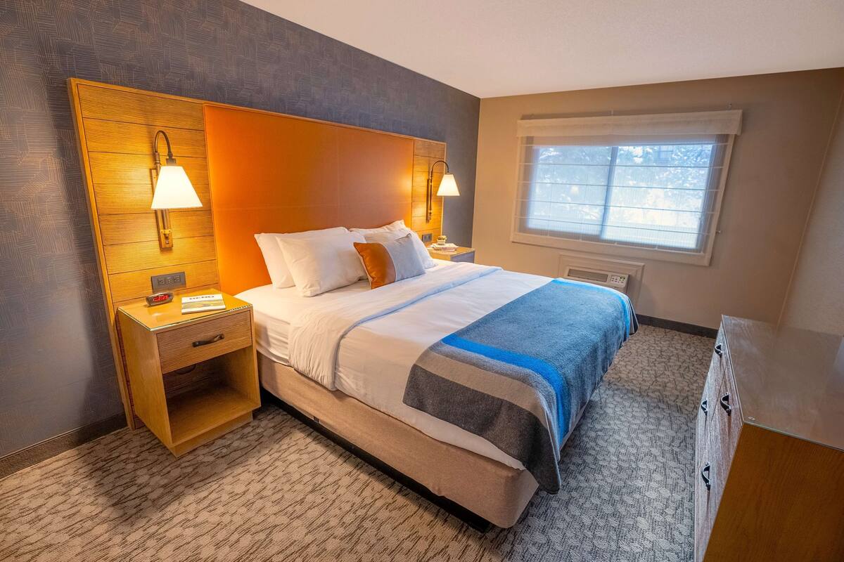 A hotel room with a large bed, two bedside tables with lamps, a nightstand, a window with blinds, and a carpeted floor.
