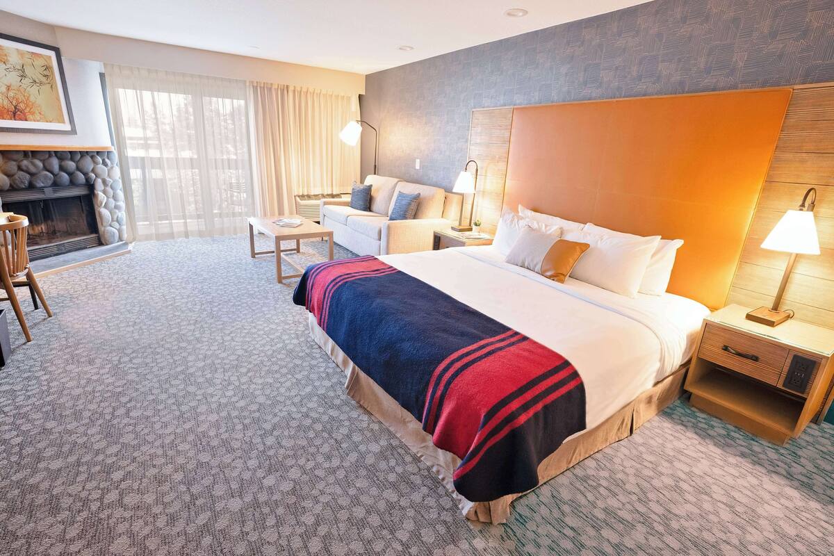 A hotel room with a large bed, a sofa, a small table, a lamp, and a fireplace.