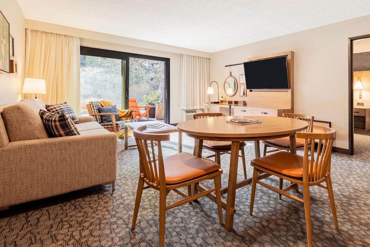 A hotel room with a sofa, a dining table with chairs, a TV, a desk, and a window with curtains.