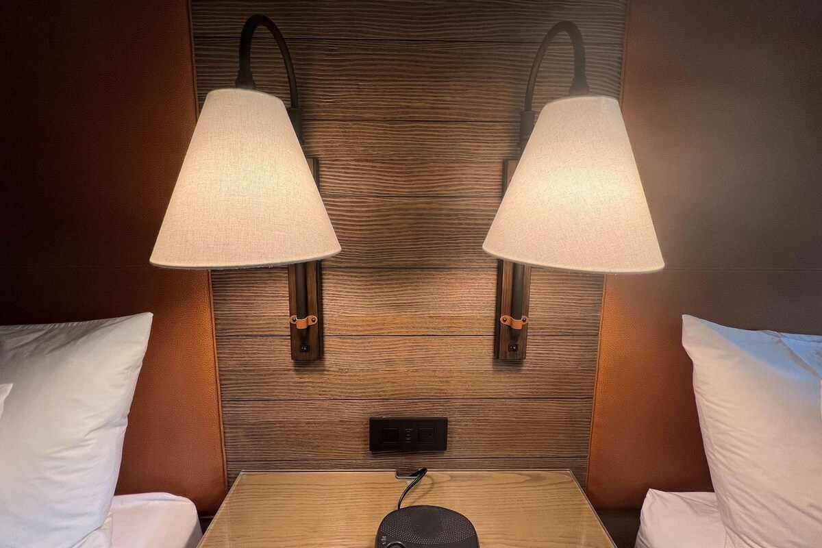 A hotel room with two wall-mounted lamps, a wooden headboard, a bedside table with a speaker, and white pillows.