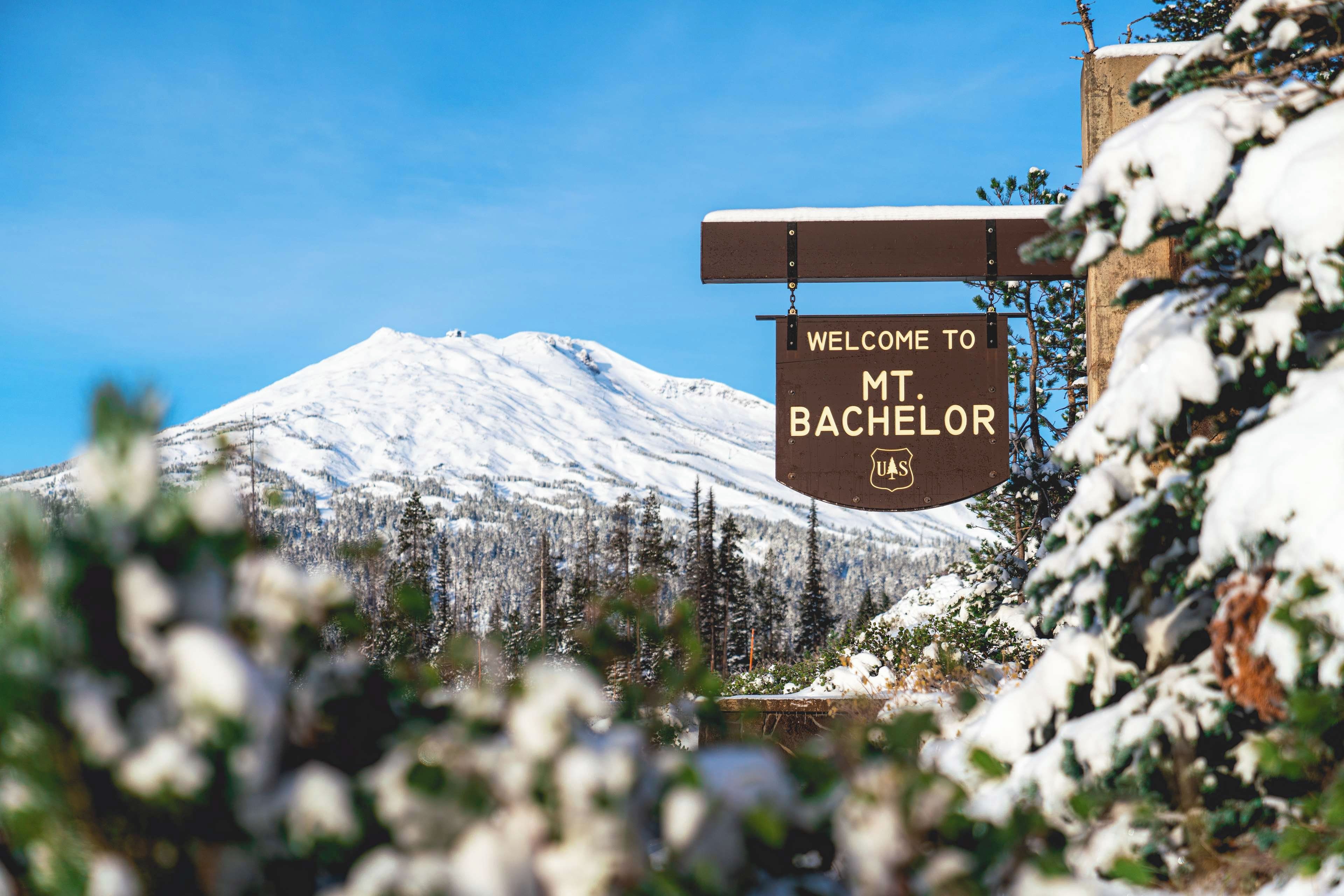 Un cartello di benvenuto per i visitatori al Monte Bachelor, con sullo sfondo una montagna innevata e alberi sempreverdi.