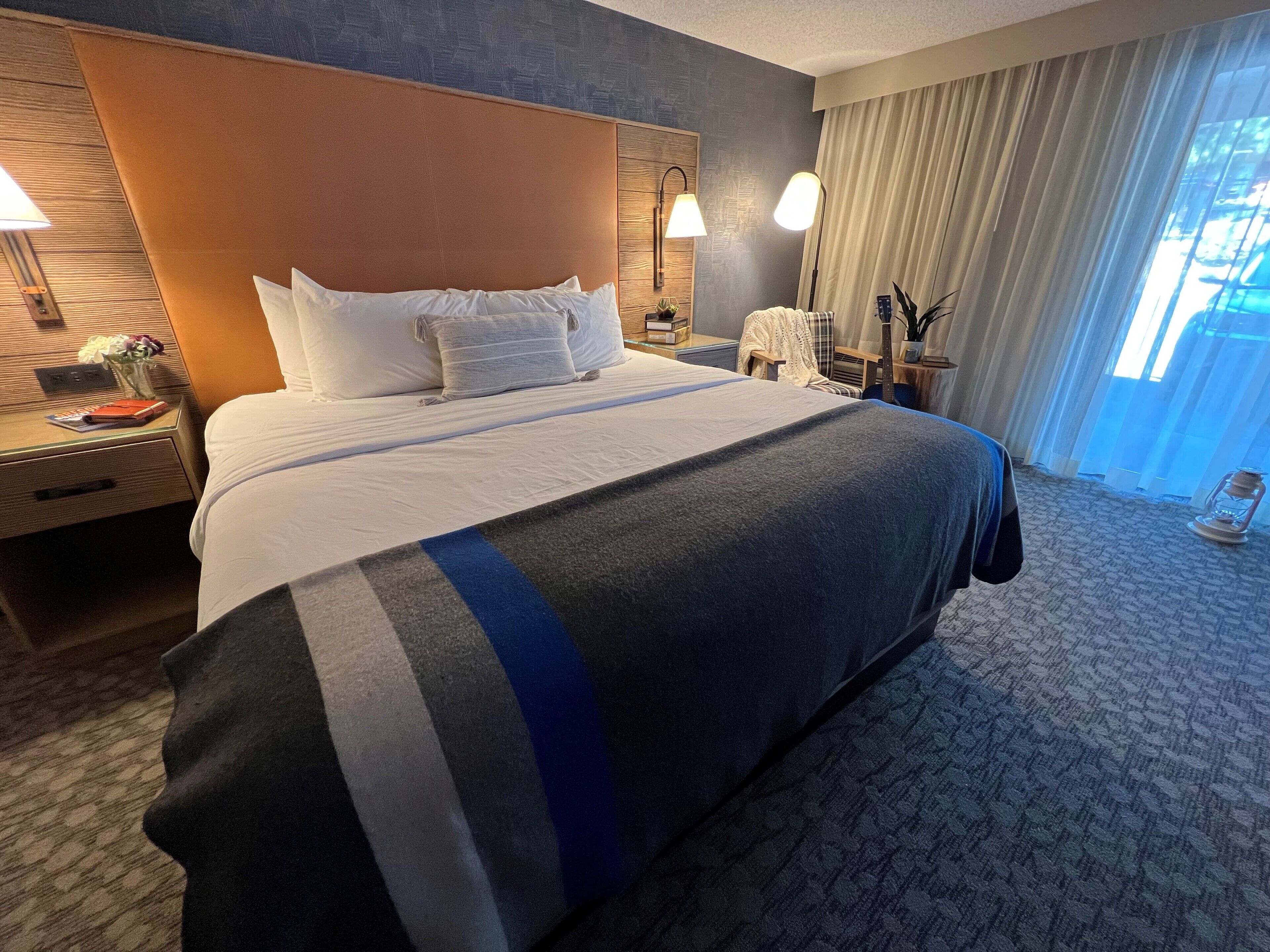 A hotel room with a large bed, a nightstand with a lamp, a window with curtains, and a view of the outdoors.