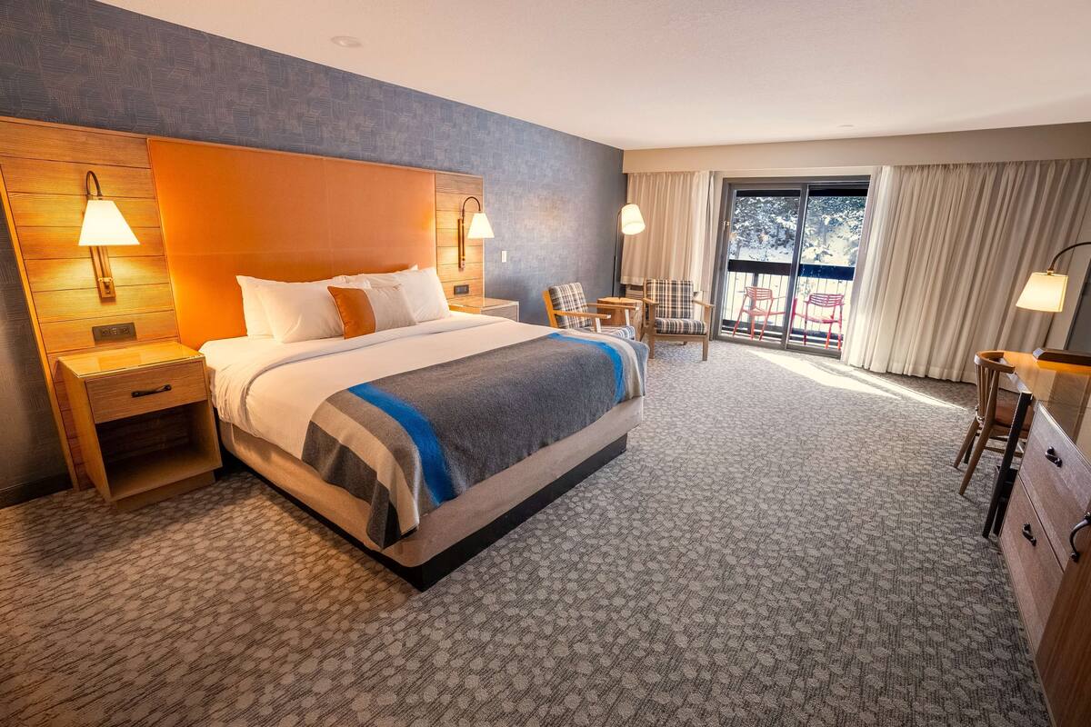 A hotel room with a large bed, two bedside lamps, a desk with a chair, and a view of the outdoors through a large window.