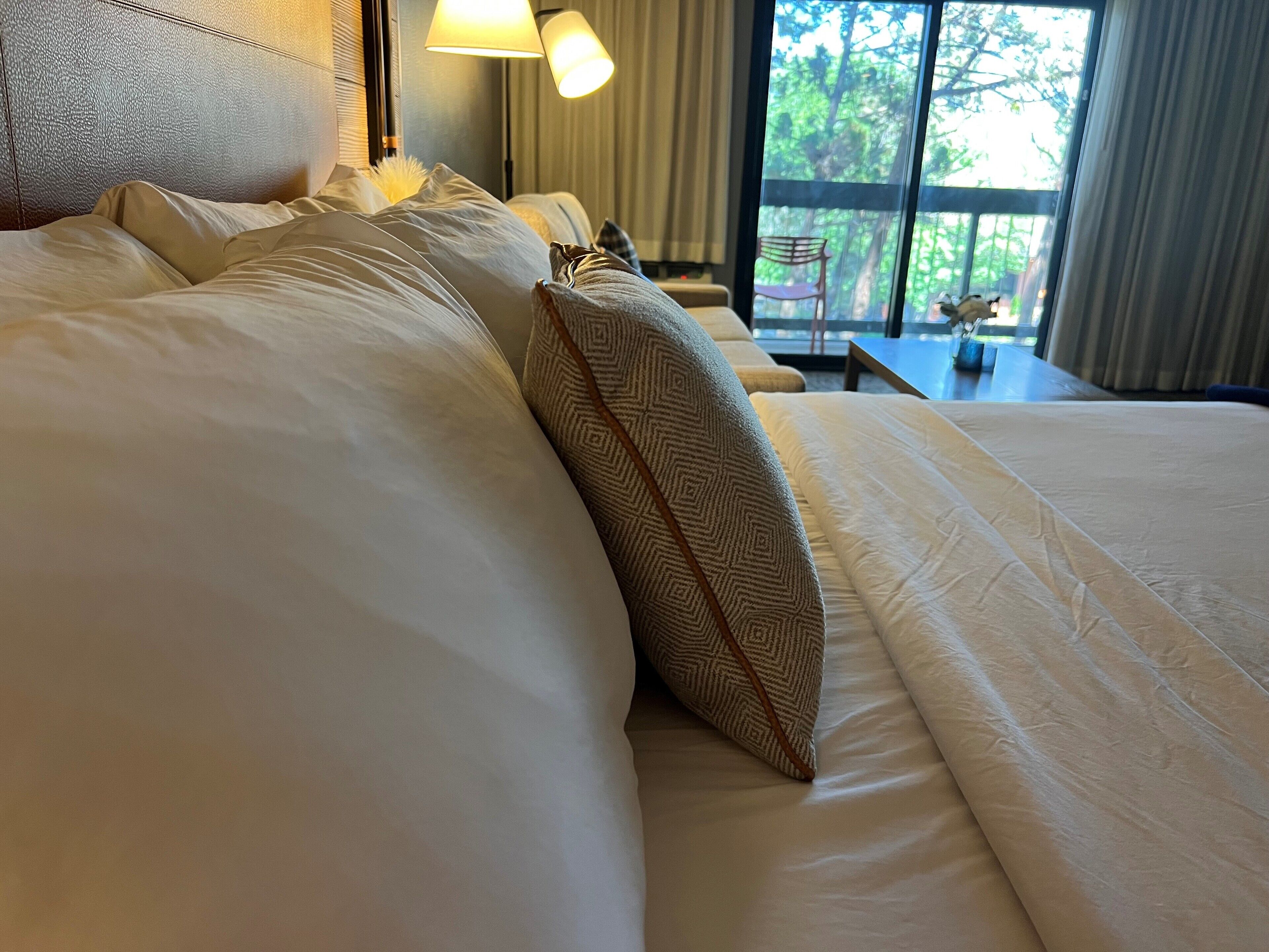 A neatly made bed with white linens and a patterned pillow, a headboard, and a lamp.