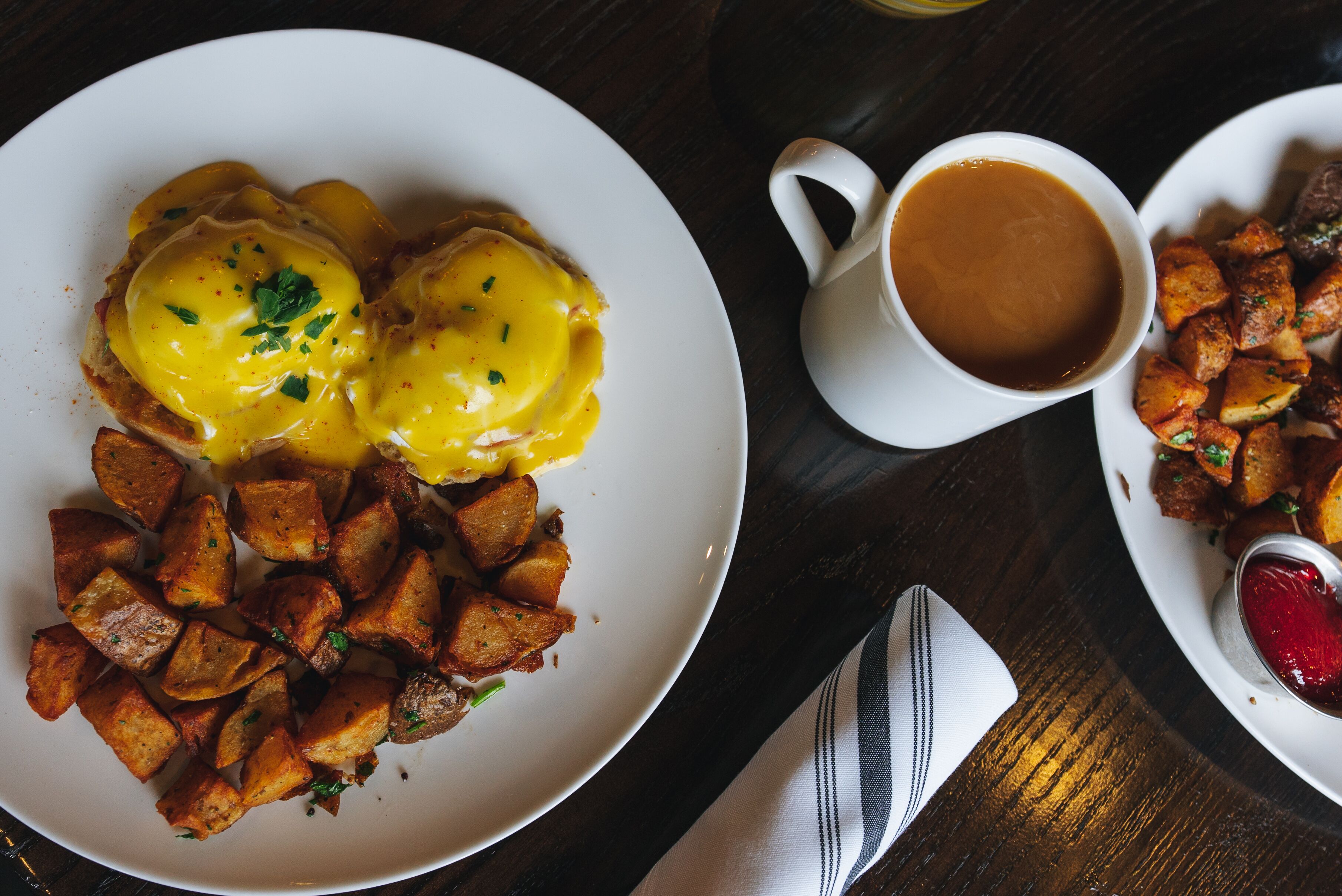 A plate of eggs benedict with hash browns and a cup of coffee on a wooden table.