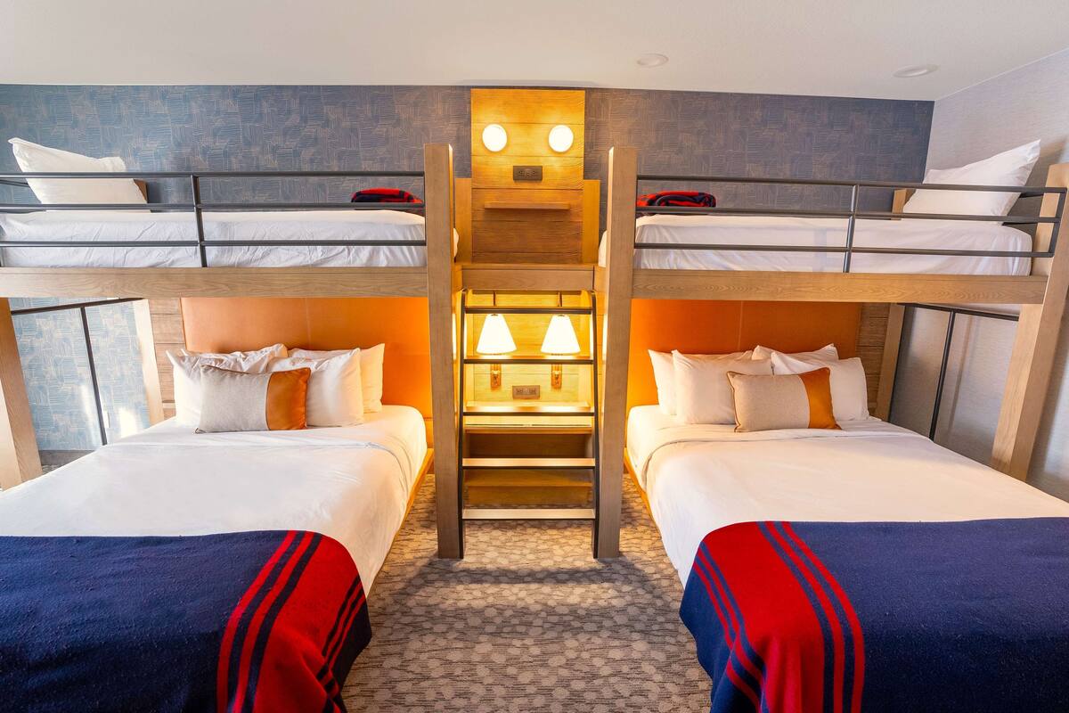 A hotel room with two single beds, a central wooden ladder, and a small table with lamps.