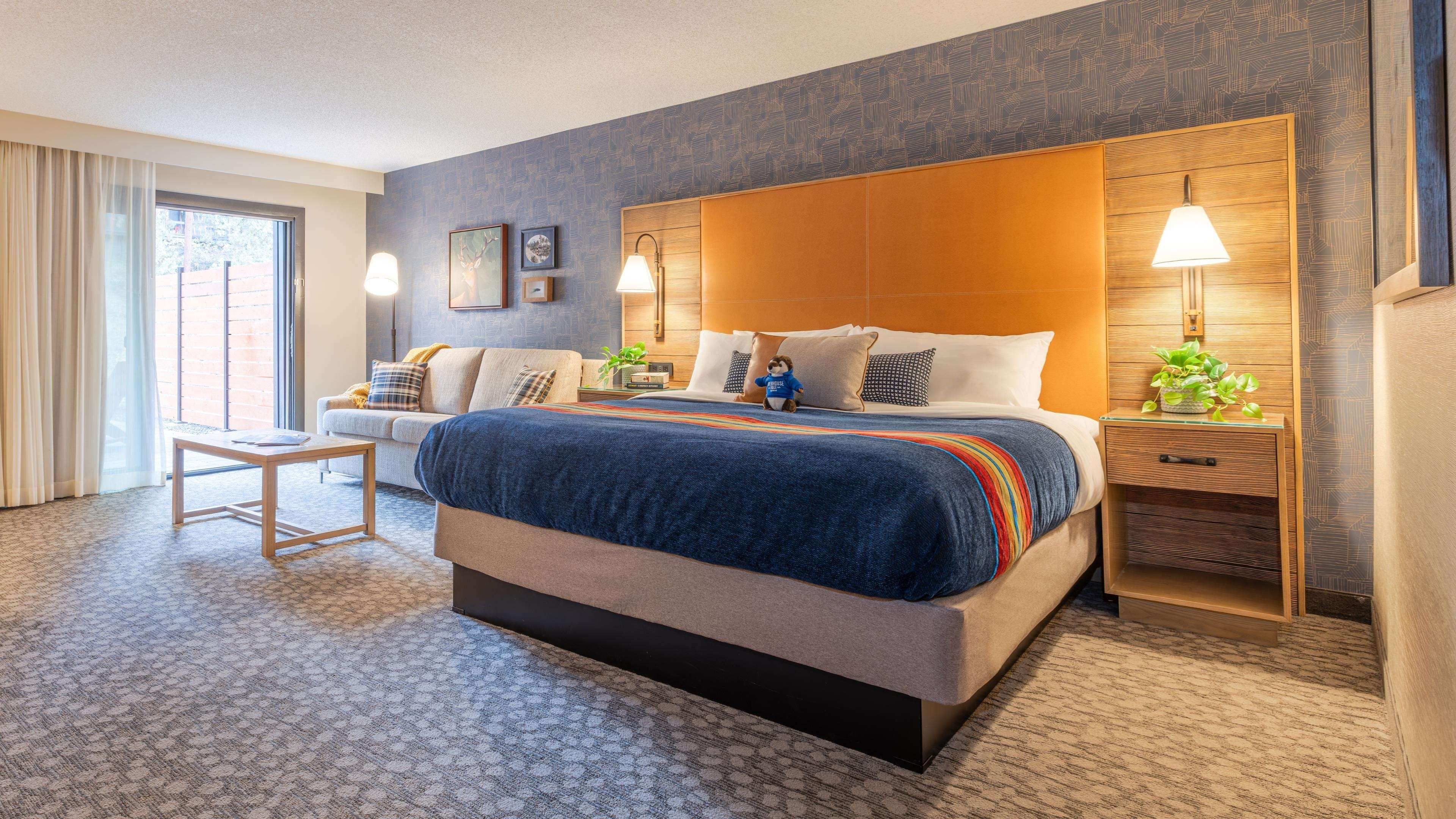 A hotel room with a large bed, a bedside table with a lamp, a small table with a chair, and a window with curtains.
