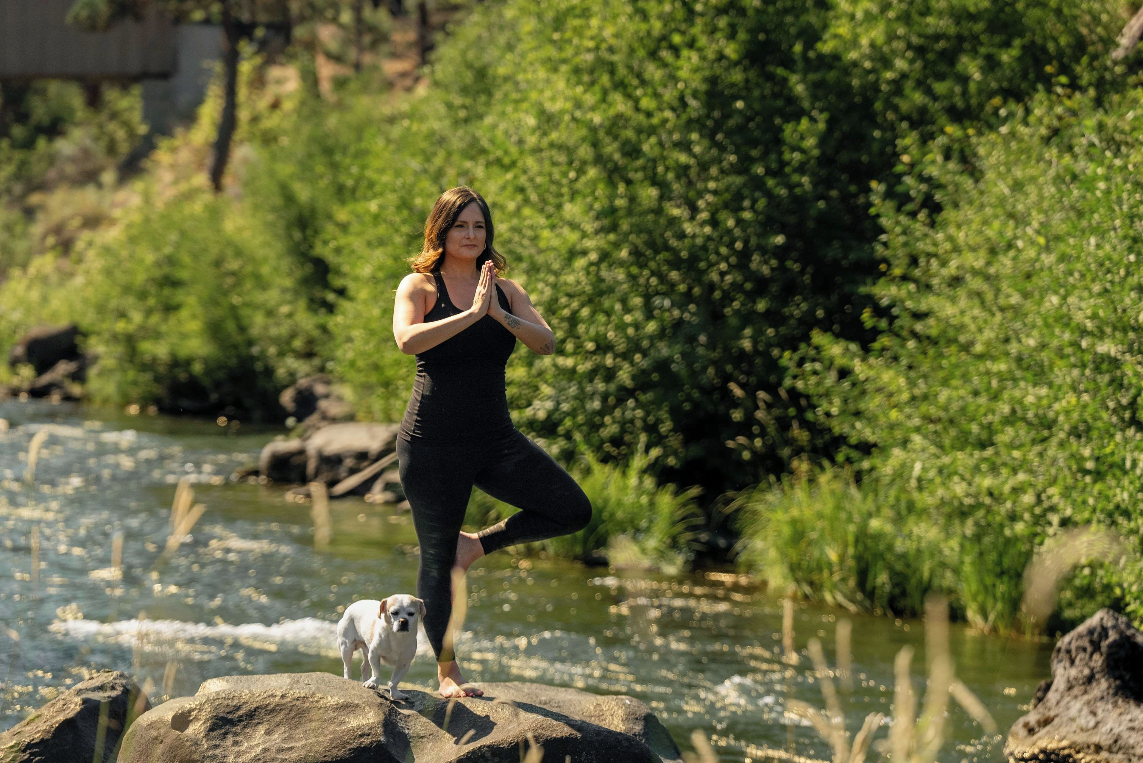 A person in a black outfit performing a yoga pose on a rock by a river with a dog nearby.