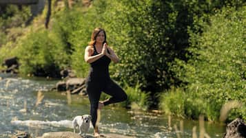 Eine Person in schwarzer Kleidung führt eine Yoga-Pose auf einem Felsen an einem Fluss aus, in der Nähe ist ein Hund.