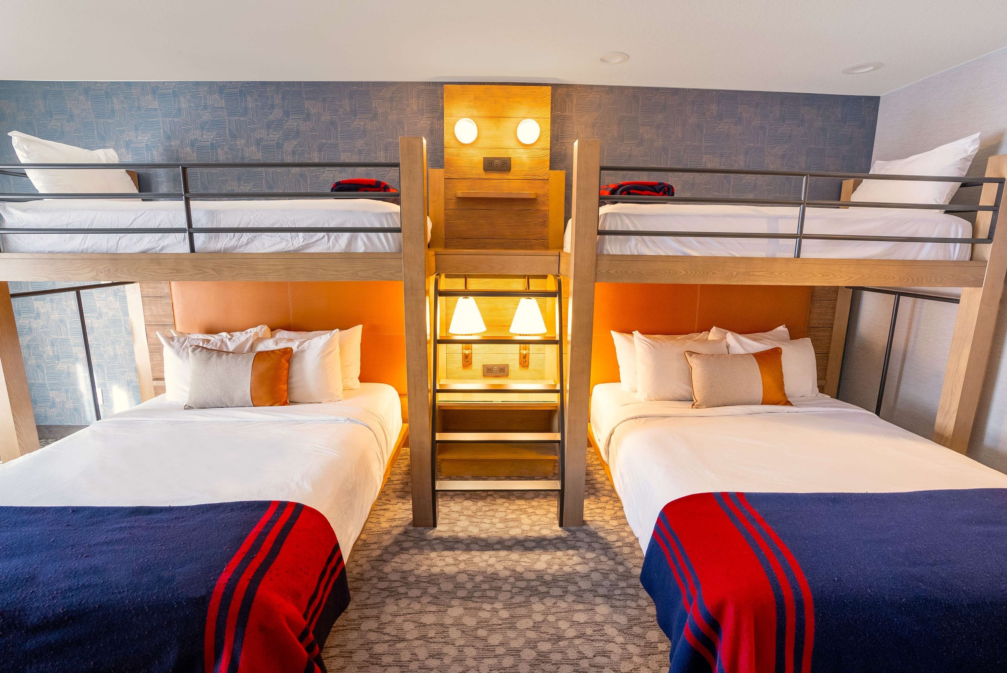 Habitación de hotel con literas, una escalera de madera central y una pequeña mesa con lámpara.