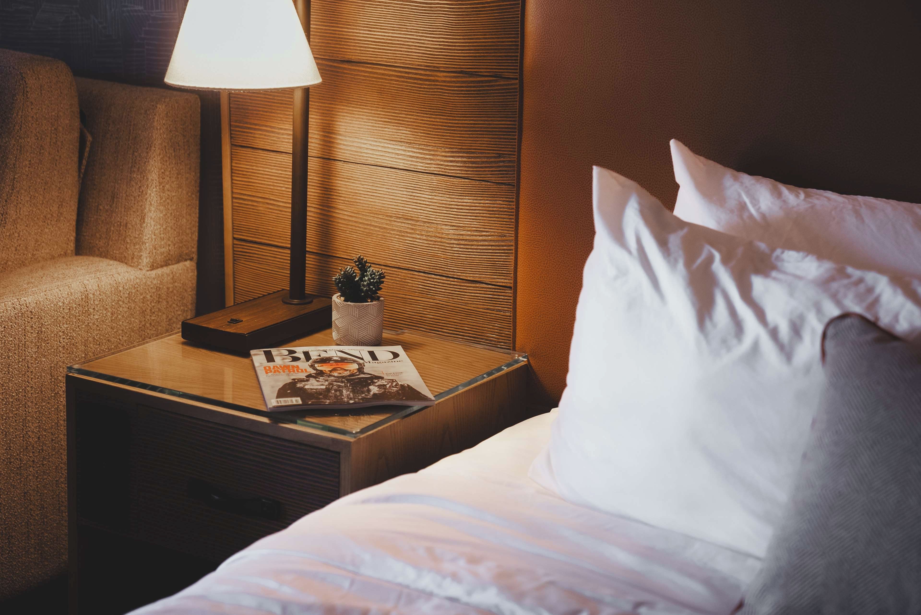 A hotel room with a neatly made bed, a bedside table with a lamp, a magazine, and a small plant.
