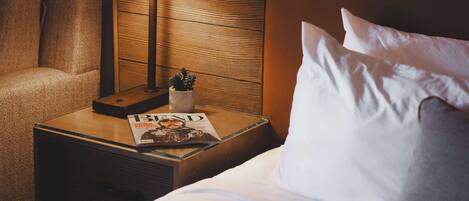 A hotel room with a neatly made bed, a bedside table with a lamp, a magazine, and a small plant.