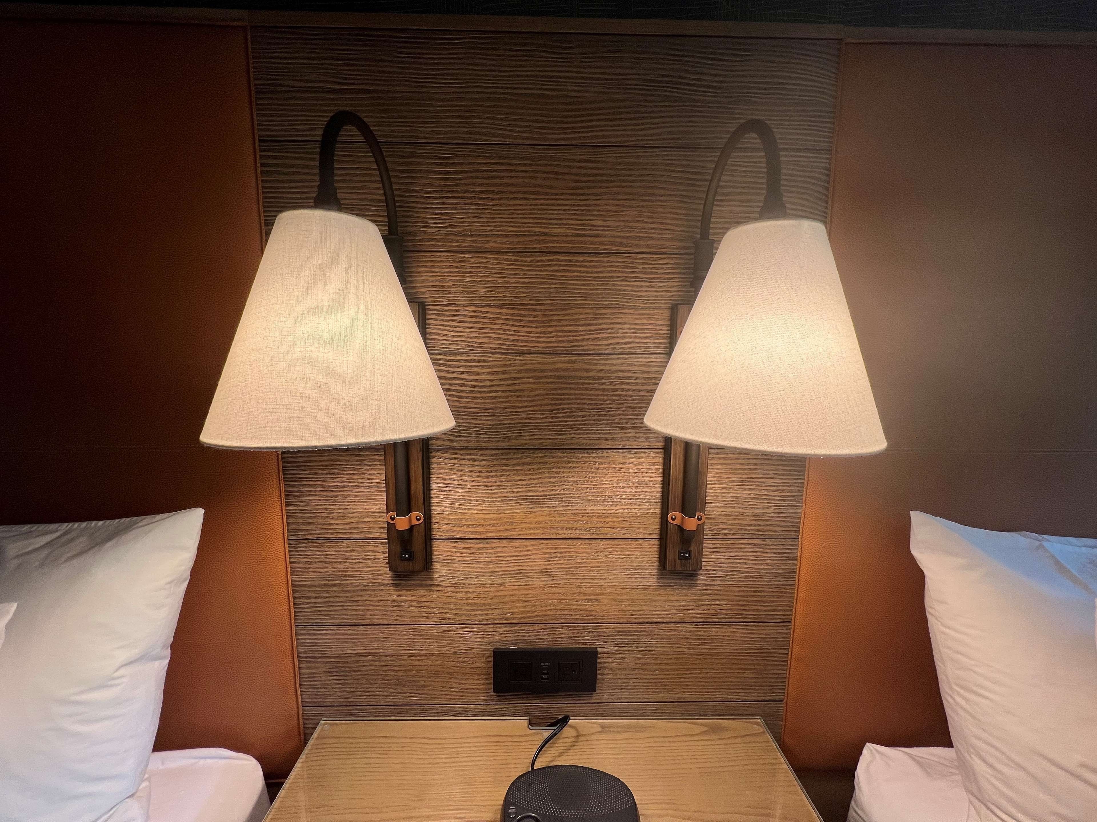 A hotel room with two wall-mounted lamps, a wooden headboard, a bedside table with a speaker, and white pillows.