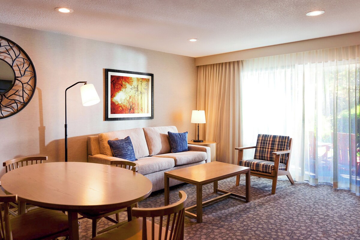 Habitación de hotel con sofá, dos sillas, una mesa, una lámpara y un ventanal con cortinas.
