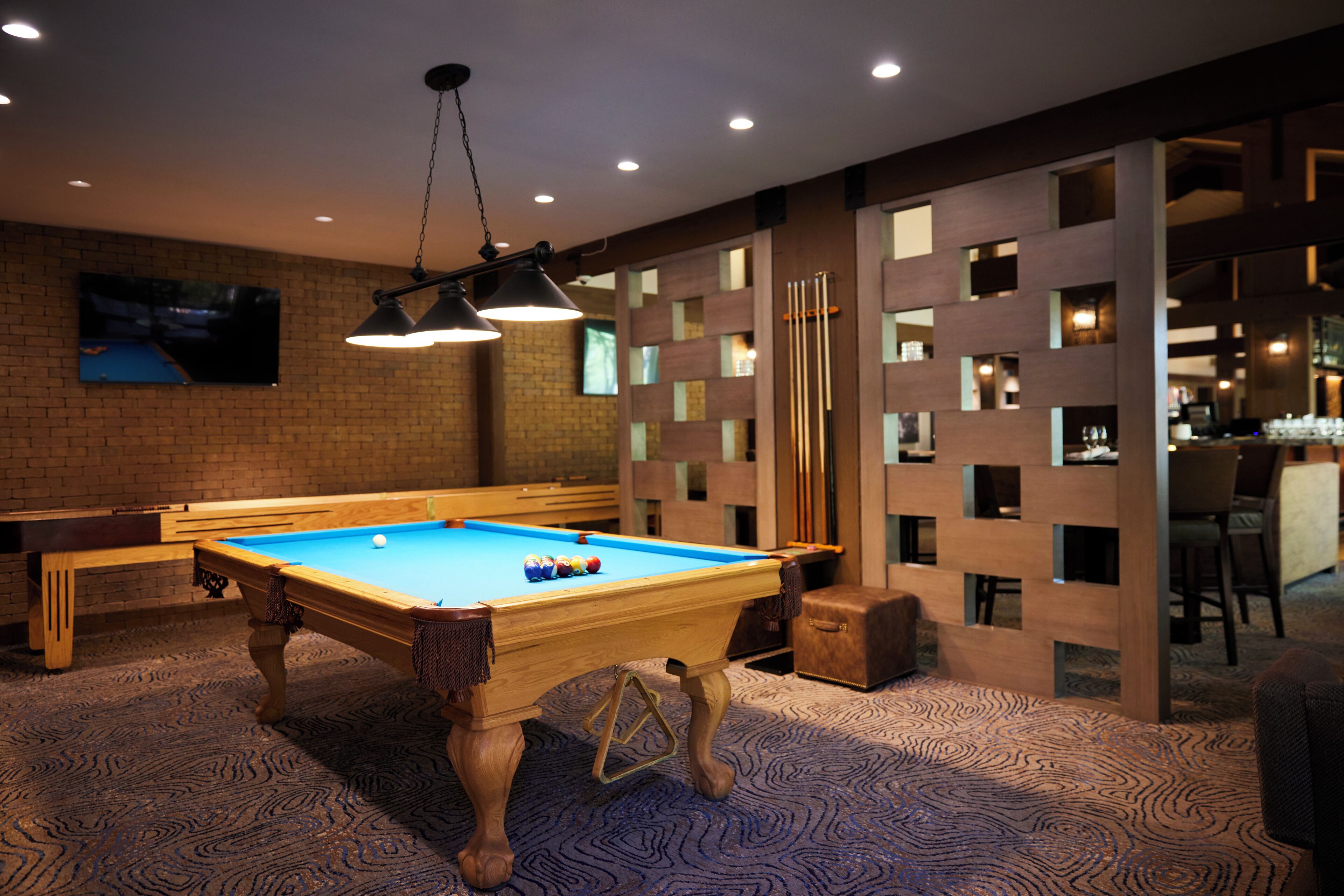 A pool table with balls, a billiard cue, and a pool table cloth. There is a television mounted on the wall and a bar area with stools.