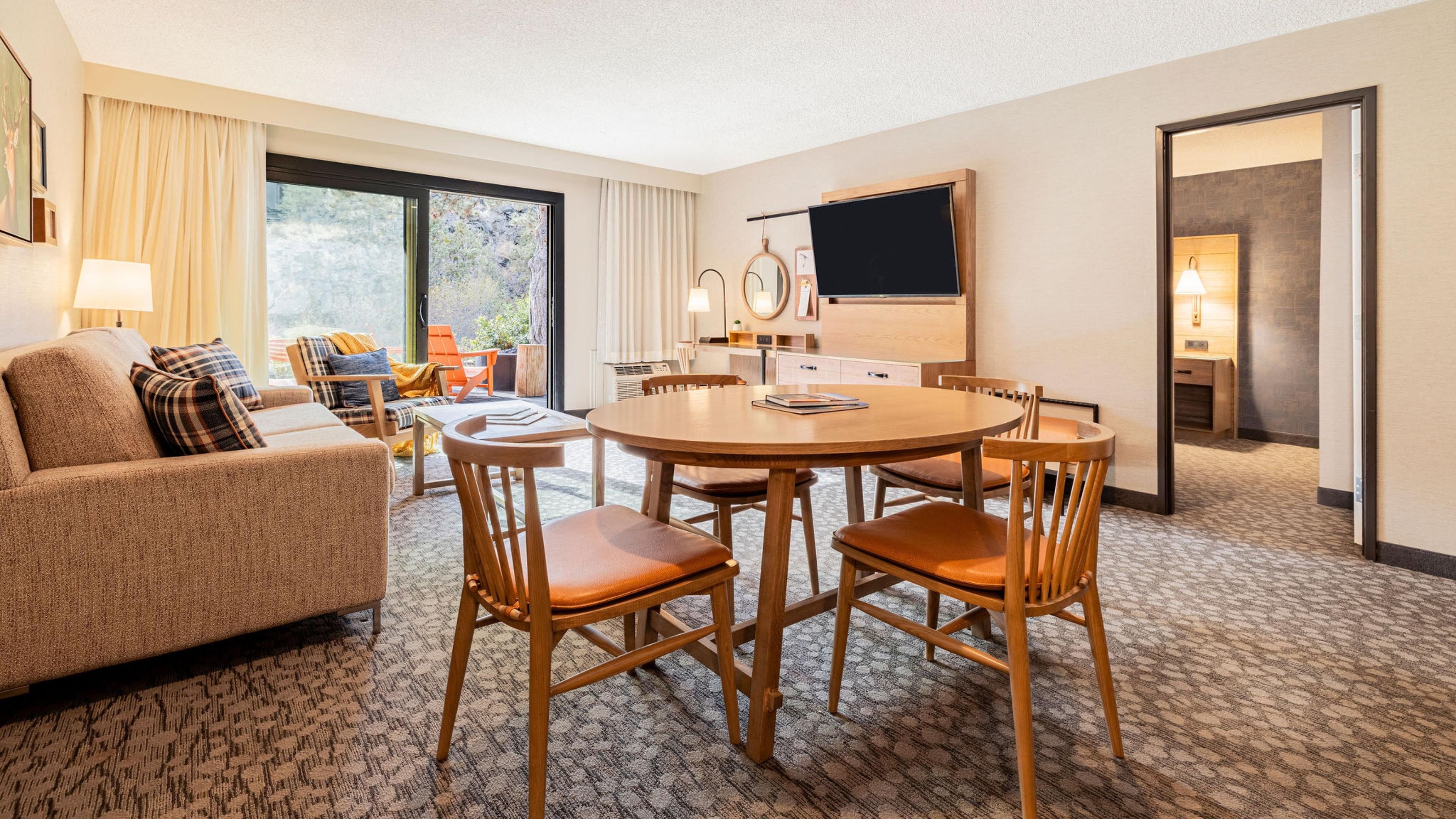 A hotel room with a sofa, a dining table with chairs, a TV, and a window with curtains.