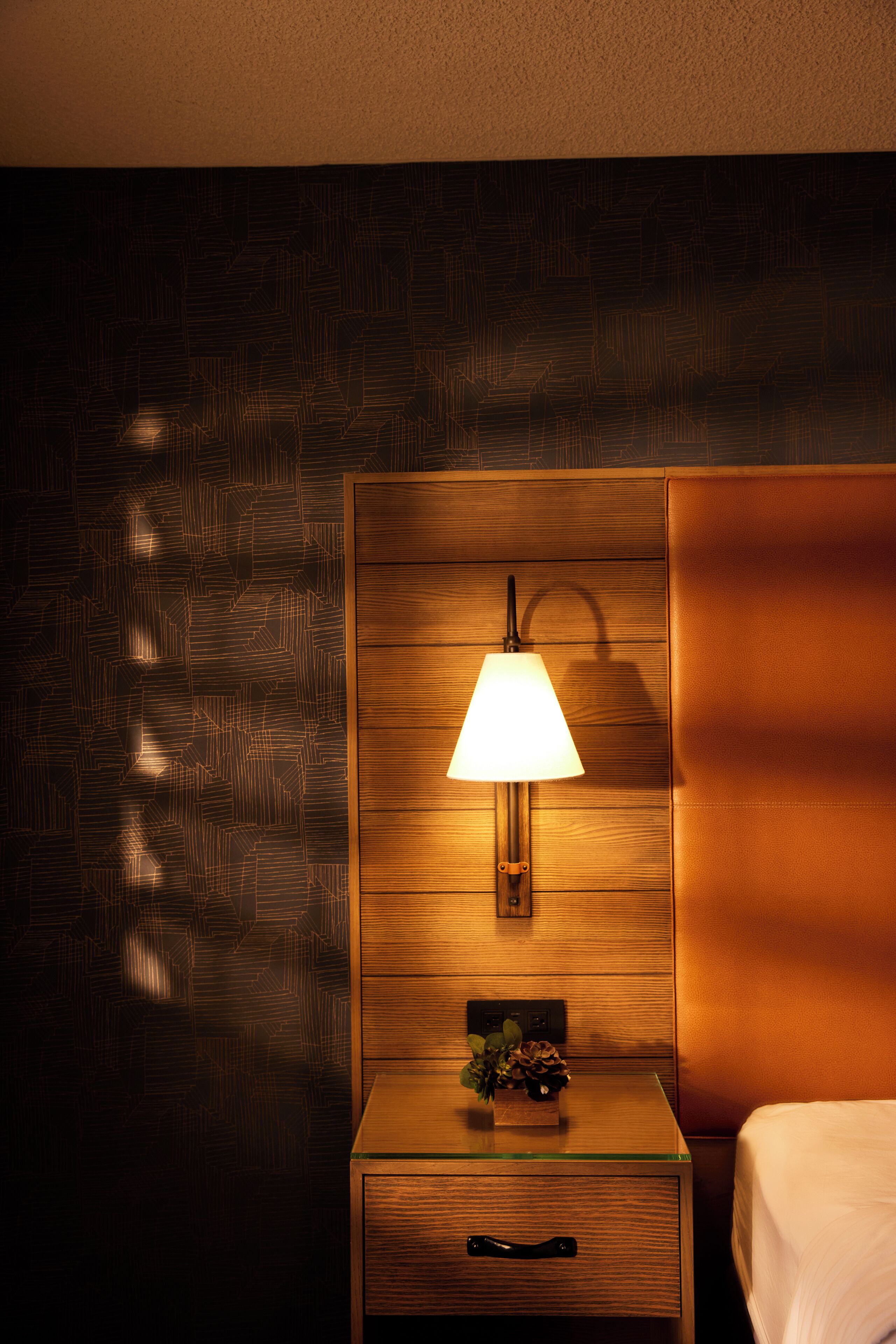 Quarto de hotel com cabeceira de madeira, luminária de cabeceira, pequena mesa com planta e cama.