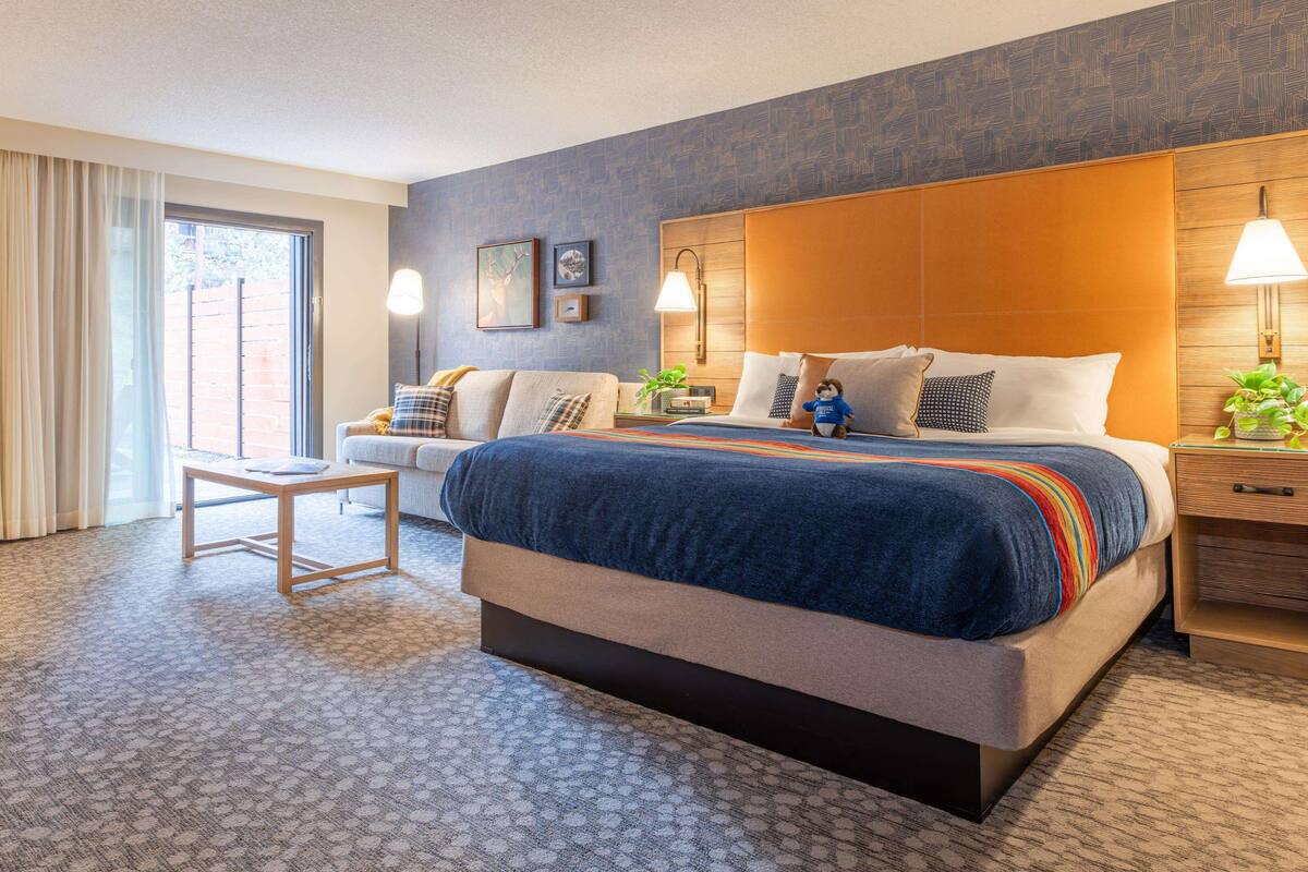 A hotel room with a large bed, a bedside table with a lamp, a small table with a chair, and a window with curtains.