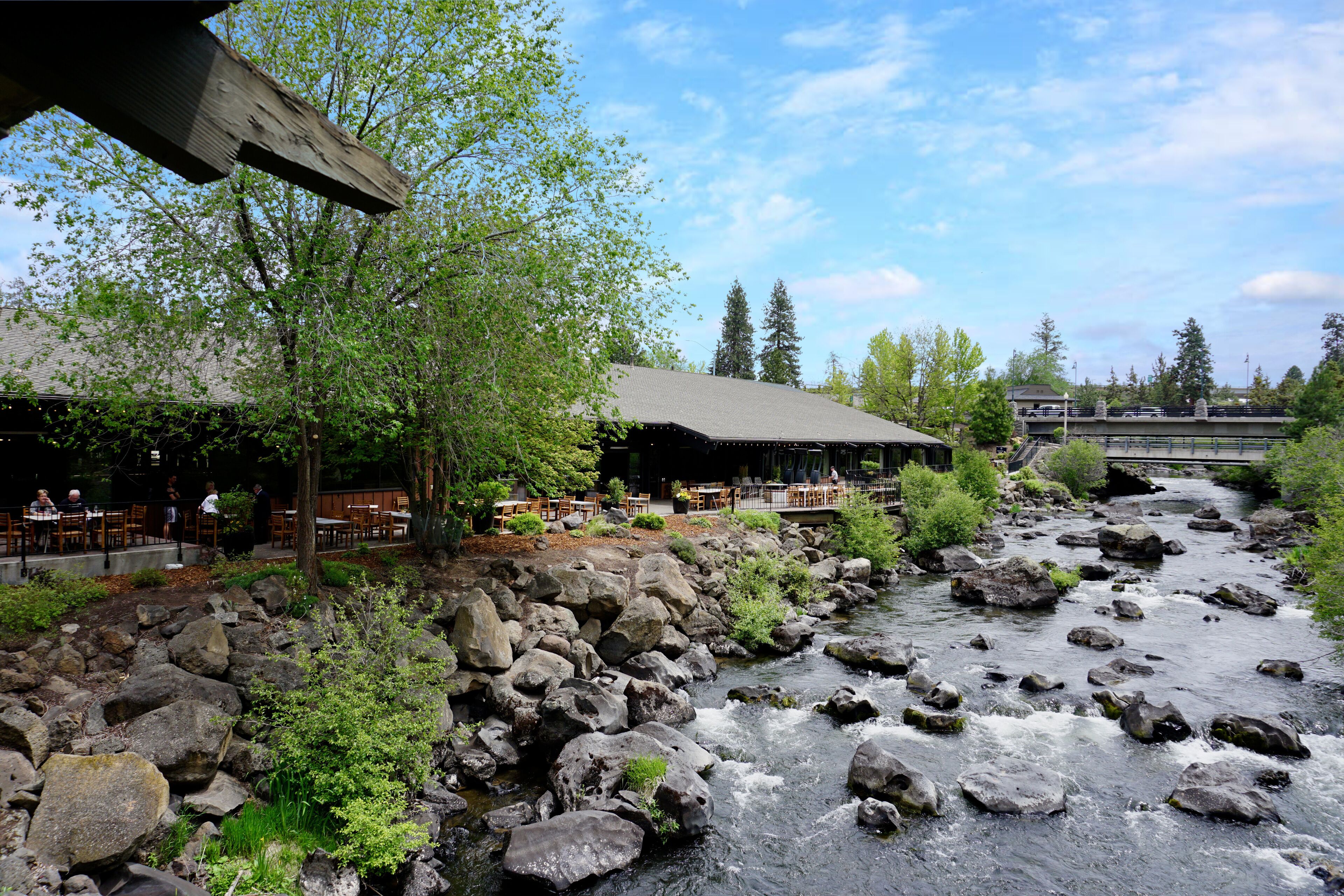 Un restaurant situé au bord de la rivière, doté d’une terrasse, d’un mur de soutènement en pierre et d’un pont en arrière-plan.