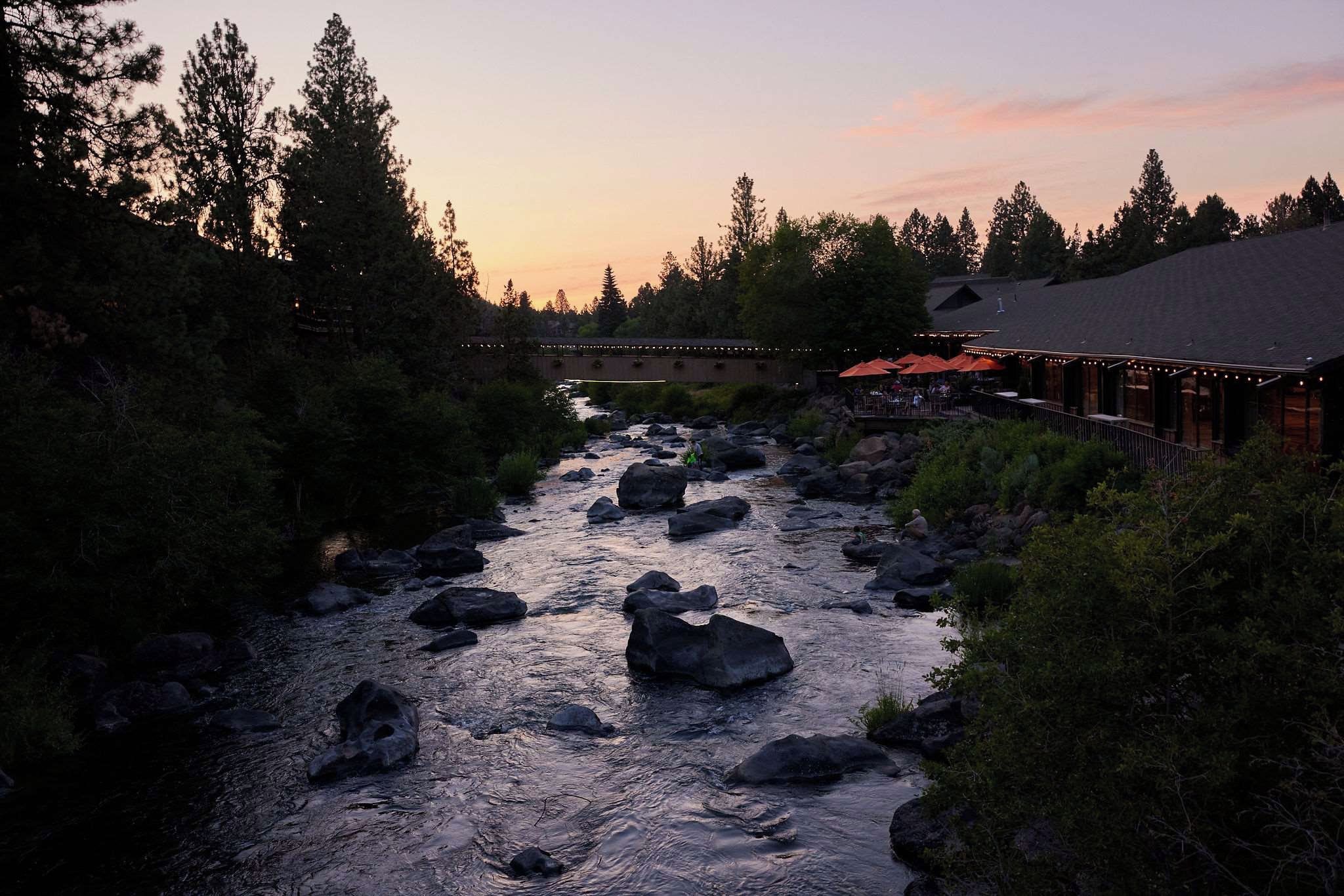 A river with rocks, a wooden bridge, and a building with outdoor seating.