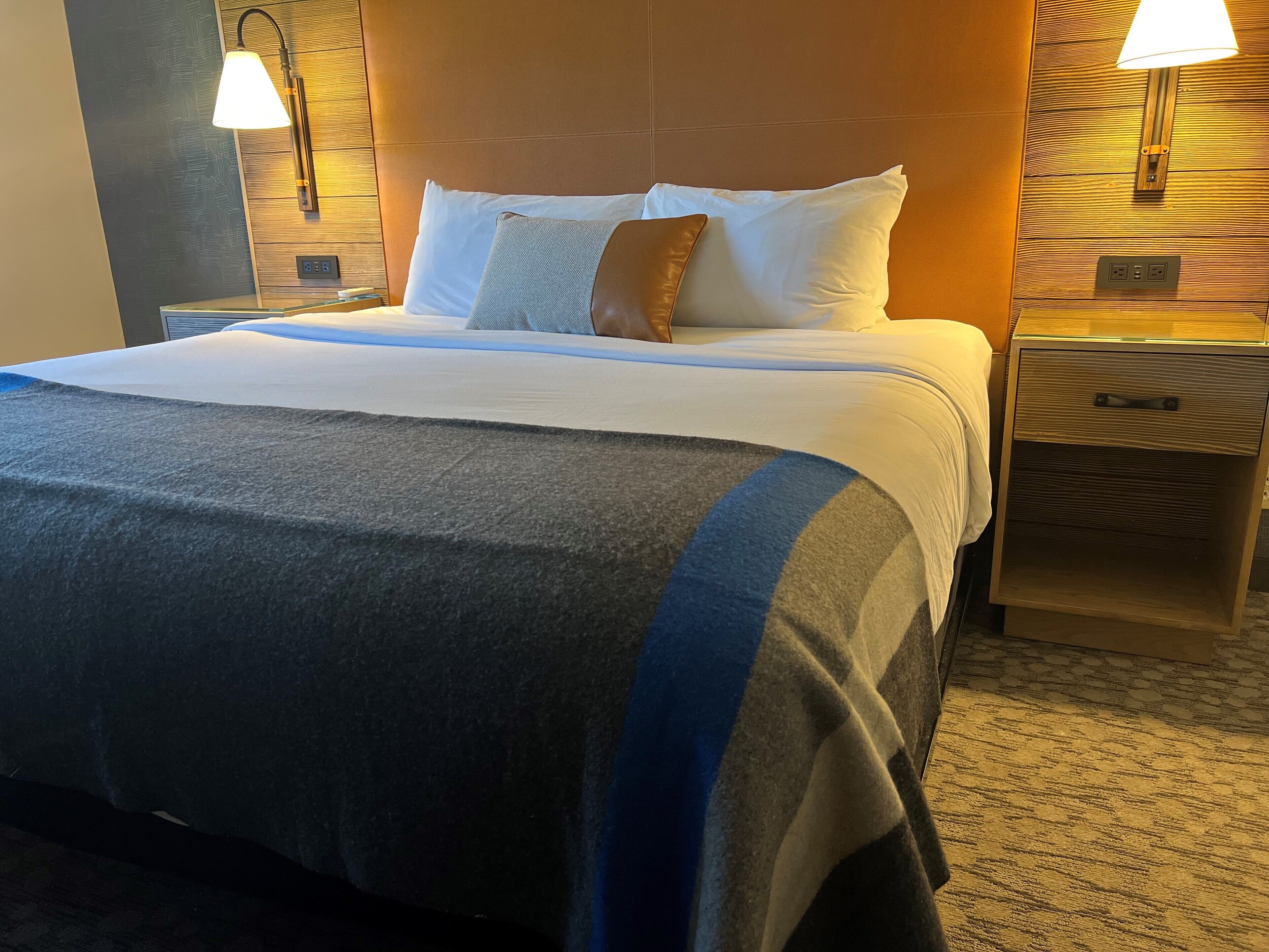 Een hotelkamer met een groot bed, twee bedlampen, een houten hoofdbord en een nachtkastje met een lade.