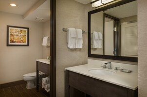 A modern bathroom with a white sink, a large mirror, and a toilet. There are towels hanging on a rack and a framed picture on the wall.