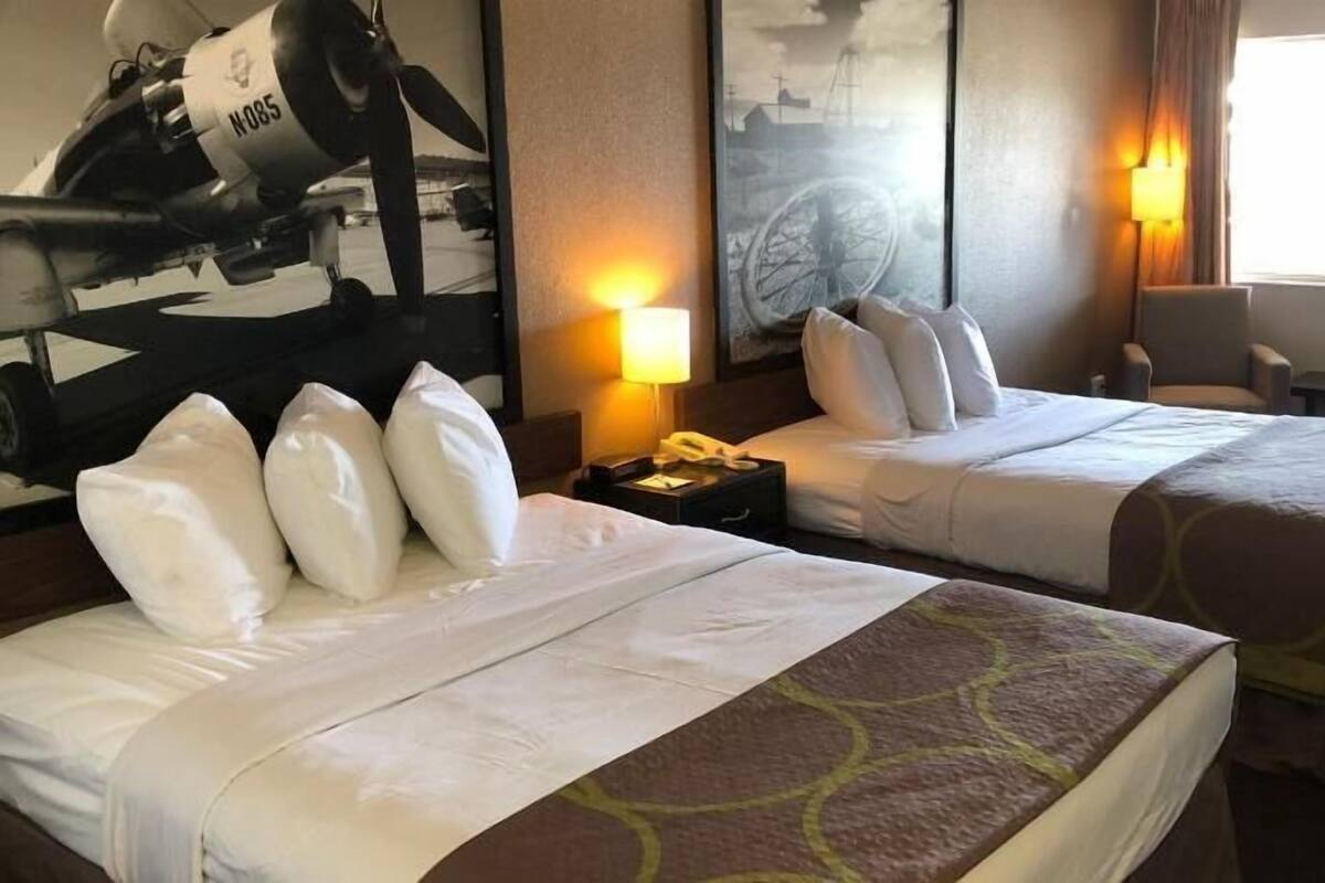 Habitación de hotel con dos camas, un escritorio, una silla, una lámpara y un cuadro de un avión.