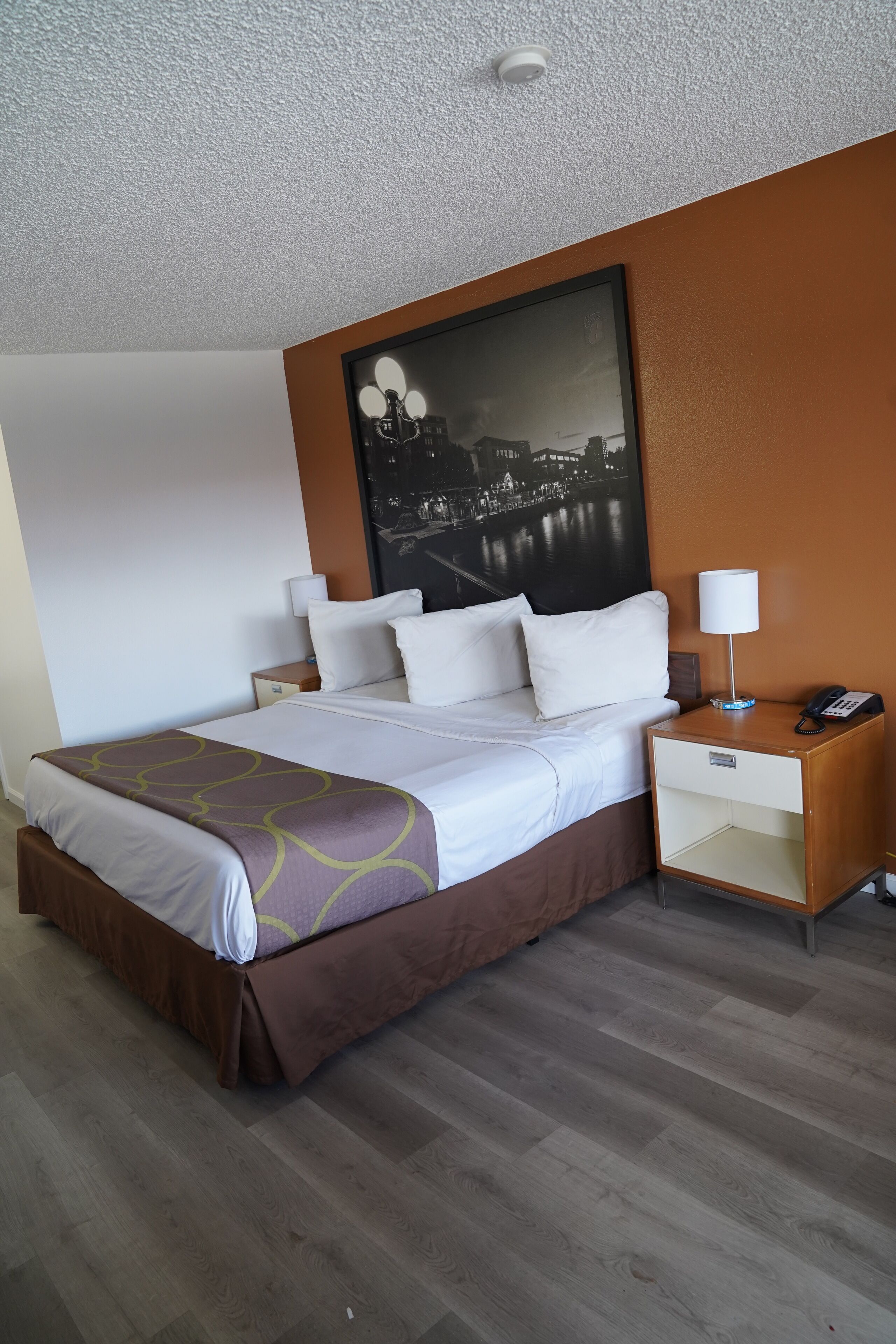 A hotel room with a bed, bedside tables, a nightstand, a lamp, and a wall-mounted artwork.