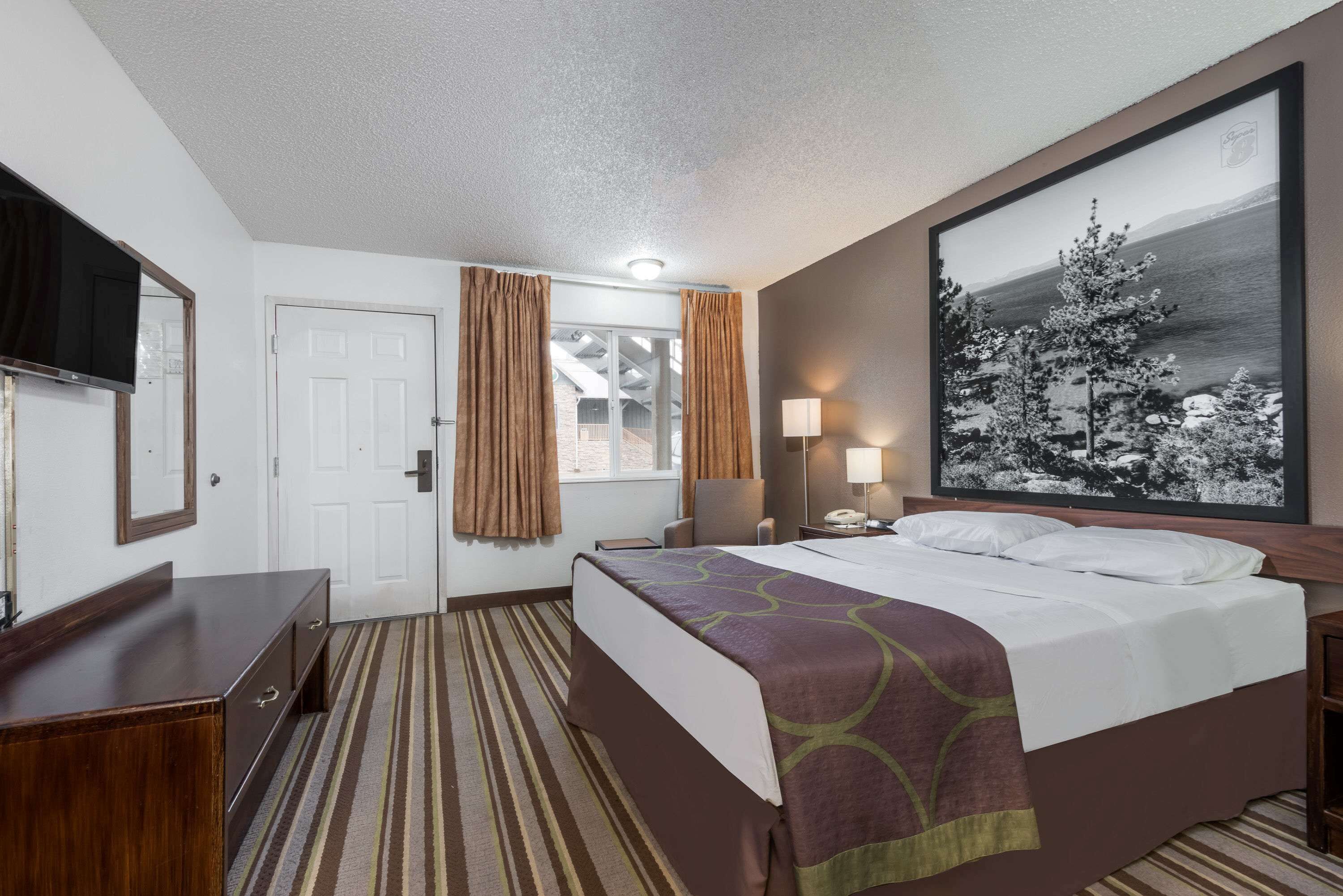 A hotel room with a bed, a desk, a chair, a TV, a window with curtains, and a painting on the wall.