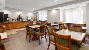 Zona pranzo dell'hotel con tavoli e sedie in legno, un bancone della cucina con macchine da caffè e un'ampia finestra con persiane.