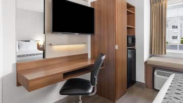 A compact hotel room with a built-in desk, a flat-screen TV, a black office chair, a microwave, and a window with curtains.