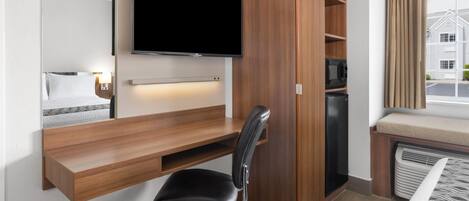 A compact hotel room with a built-in desk, a flat-screen TV, a black office chair, a microwave, and a window with curtains.
