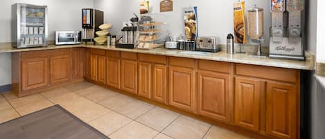 Ein Frühstücksbereich eines Hotels mit Selbstbedienungsstation, einer Kaffeemaschine und einem Kühlschrank mit Lebensmitteln.
