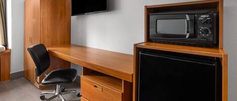 Quarto compacto com uma escrivaninha de madeira, uma cadeira de escritório preta, televisão fixada na parede e micro-ondas em uma prateleira de madeira.