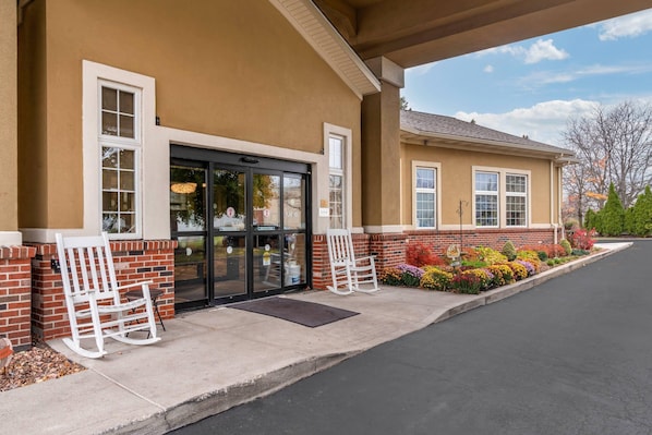 A building with a beige exterior, a black entrance door, and a brick lower section. There are two white rocking chairs outside.
