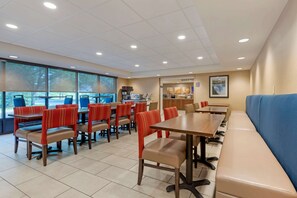 A dining area with multiple tables and chairs, a blue bench, and large windows with a view of trees.