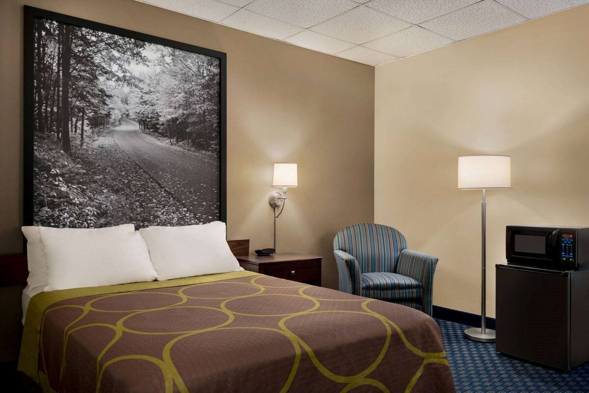 A hotel room with a large bed, a striped armchair, a desk with a lamp, and a refrigerator.