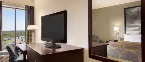 A hotel room with a desk, chair, television, bed, and a large window offering a view of the city.