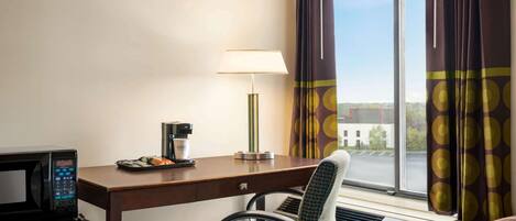 A hotel room with a desk, a chair, a microwave, a coffee maker, and a window with curtains.