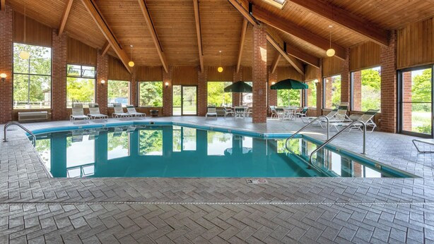 An indoor swimming pool with a wooden ceiling, brick walls, and large windows. - Baymont by Wyndham Harvard (Harvard)