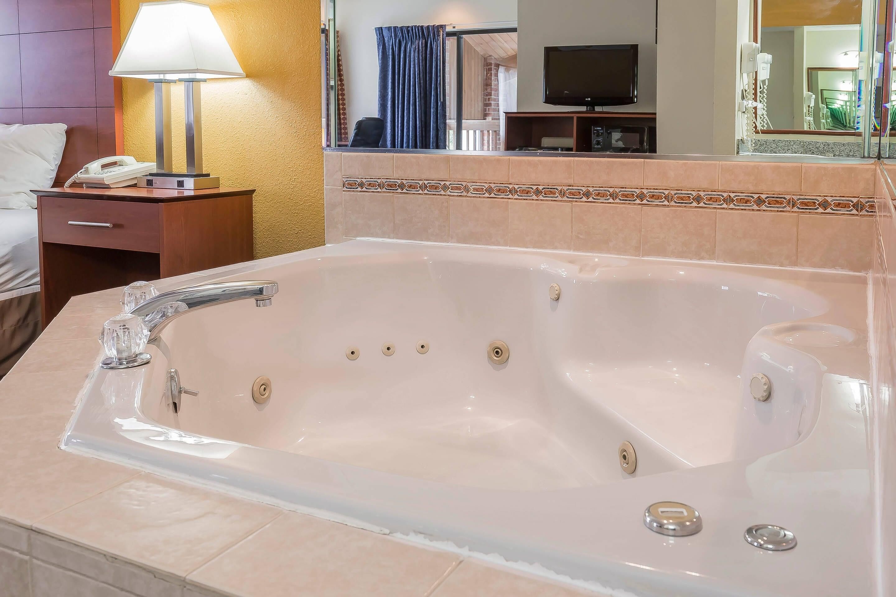 A hotel room with a large bathtub, a bedside table with a lamp, a TV, and a mirror.