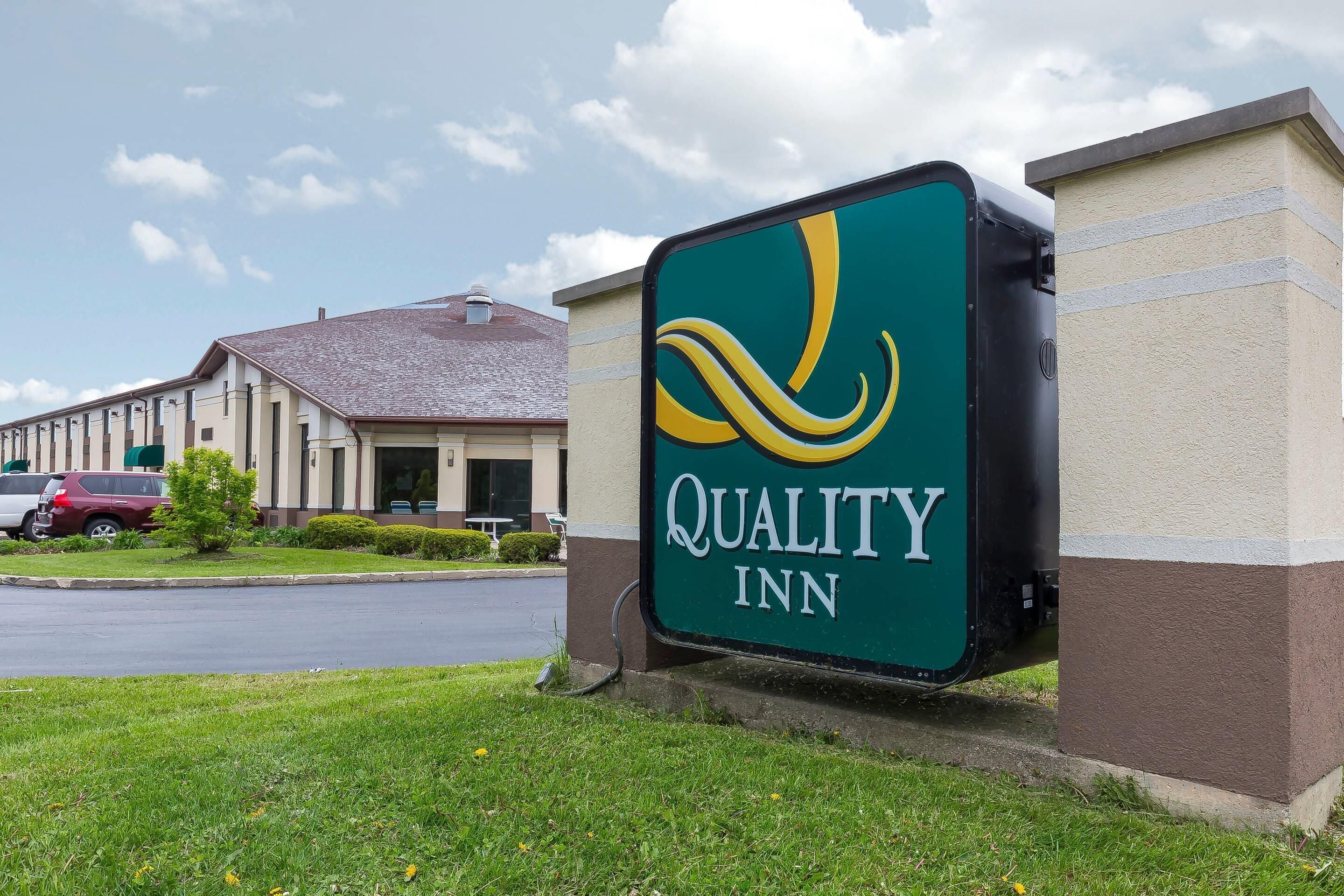 A Quality Inn sign with a green and yellow logo, a building with a brown roof, and a parking lot with cars.